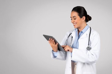 Beyaz üniformalı neşeli Latin kadın doktor dijital tablet kullanıyor. Cihazla gülümseyen doktor hastalara internet üzerinden danışmanlık yapıyor. Gri arkaplanda poz veriyor.