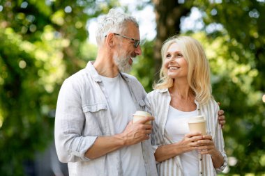 Emeklilik. Gülümseyen kıdemli eşler dışarıda yürüyor ve paket kahve içiyor, mutlu olgun erkek ve kadın parkta yürüyor, romantik yaşlı çift dışarıda birlikte dinleniyor, boş alan var.