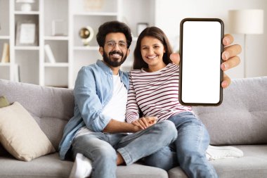 Sevimli, neşeli, mutlu, binlerce yıllık Hintli çift rahat bir oturma odasında kanepede oturuyor, sarılıyor ve gülümsüyor, akıllı telefonları beyaz ekranla birlikte evde online alışveriş yapıyorlar.