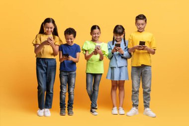 Çocuklarda Gadget bağımlılığı. Sarı arka planda izole edilmiş akıllı telefonları kullanan çeşitli ergen çocuklar. Mobil oyunlar oynayan, kaydıran, çevrimiçi içeriği izleyen çok ırklı okul çocukları ve kızları