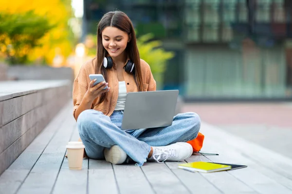 スタイリッシュなカジュアルな服装大学の学生で陽気で美しい若い女性がプロジェクトに取り組んでいます 都市部の階段に座って 屋外のラップトップコンピュータを使用して スマートフォンをチェック 無料スペース — ストック写真