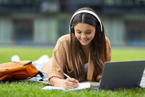 スマートな美しい若い女性大学の学生は公園で冷笑し 単独で芝生に横たわり ワイヤレスヘッドフォンとラップトップコンピュータを使用して ノートを取るか ウェビナーに出席するか オンラインレッスンを持っています 教育について — ストック写真