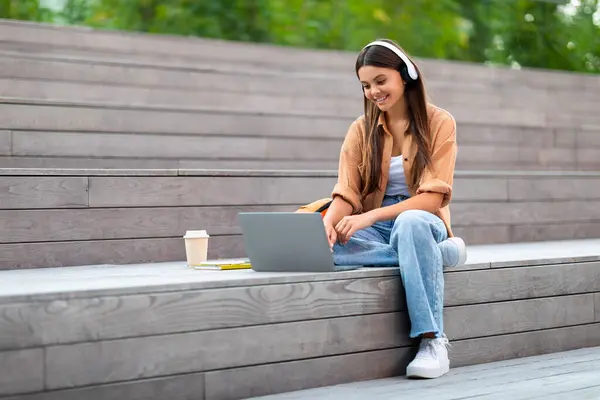 在城市地区的楼梯上坐着一位非常时髦的年轻女性嬉皮士 她用的是电脑笔记本电脑 户外无线耳机 外卖咖啡 看网络研讨会 做作业 空空间 — 图库照片