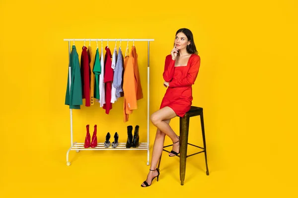 What To Wear. Thoughtful Woman Sitting On Chair Near Rack With Clothes And Shoes, Pensive Young Shopaholic Lady Cant Decide How To Dress, Posing Over Yellow Studio Background, Copy Space