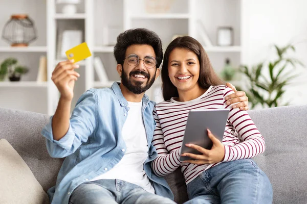 Evlerinde dijital tablet ve banka kartı kullanan neşeli, genç Hintli çift. Kanepede oturan mutlu adam ve kadın, alışveriş veya bankacılık, mal ve hizmetler için ödeme, kopyalama alanı.