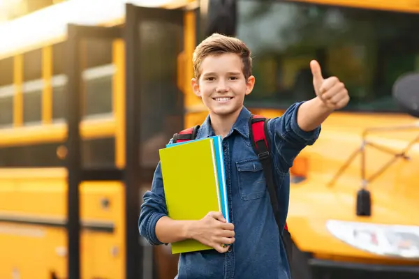 微笑着站在黄色校车旁边的十多岁男孩 并在相机前展示自己的照片 快乐的男孩手握课本 享受学习 推荐教育计划 复制空间 — 图库照片