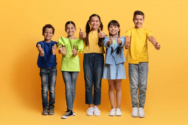 stock image Cute cheerful muliethnic school aged kids posing isolated on yellow background, showing thumb ups and smiling at camera. Cute preteen boys and girls having fun together, recommending nice kids club