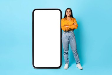 Başvuru Reklamı. Büyük Cep Telefonu 'nun yanında boş ekranı olan genç Hintli kadın Mavi Arkaplan' ın üzerinde akıllı telefon uygulaması için kendine güvenle el ele tutuşuyor. Model