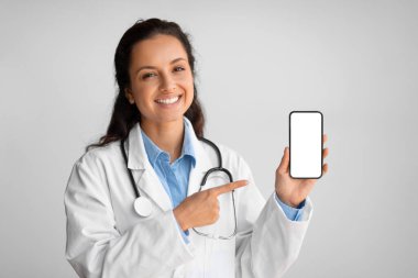 Üniformalı gülümseyen kadın doktor boş cep telefonu ekranını gösteriyor ve işaret ediyor, elinde aleti tutuyor, açık gri duvar arka planında duruyor, maket yapıyor. Teletıp kavramı