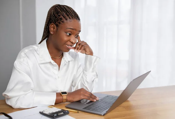 Surprised pretty young african american lady wearing formal outfit employee sitting at desk at office, reading emails on laptop, looking at screen with astonished face expression, copy space