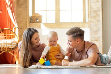 Mutlu genç ebeveynler ve onların sevimli bebekleri evde oyuncaklarla oynuyorlar, Milenyum annesi ve babası yeni yürümeye başlayan çocuğuyla birlikte oturma odasında dinlenirken eğleniyorlar,