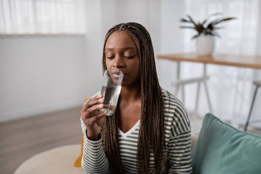 Susuzluk, sağlıklı alışkanlıklar konsepti. Uzun rastalı güzel siyahi bir bayan, günlük kıyafetler giyiyor, elinde bir bardak taze kaynak suyu tutuyor, evdeki kanepede oturuyor, reklam için fotokopi çekiyor.