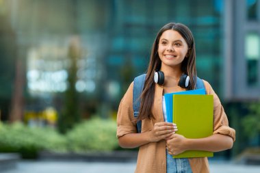 Üniversitede ya da kampüste poz veren, sırt çantası ve ders kitapları taşıyan, fotokopi alanına bakan ve gülümseyen uzun saçlı mutlu genç bayan öğrenci. Eğitim kavramı