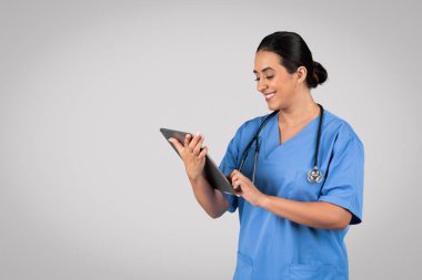 Mavi üniformalı Latin hemşirenin tablet yazmasına sevindim. Teknolojiyle çalışıyor. Gri stüdyo arka planında izole edilmiş poz veriyor. İlaç ve sağlık hizmetleri, yardım ve uzaktan muayene, çalışma
