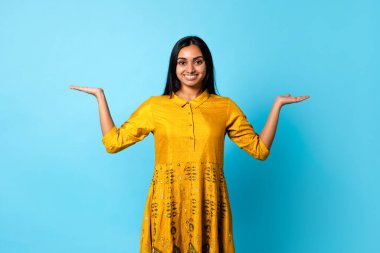 Genç Hintli bayan terazi yapıyor, seçenekleri karşılaştırıyor, mavi stüdyo arka planında görünmez nesneleri tutuyor, kameraya gülümsüyor. Orta Doğulu kadın reklam ürünleri, afiş tasarımı