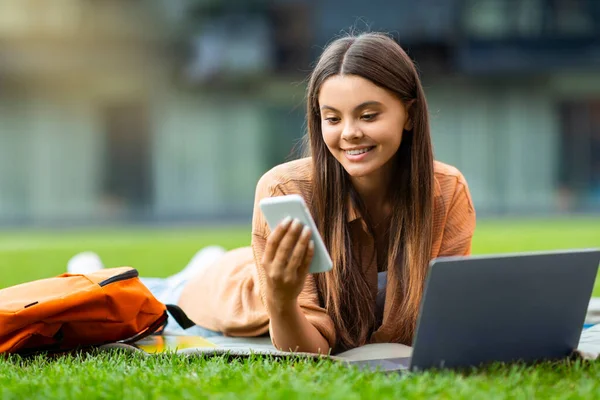 漂亮可爱的女大学生在公园里用小玩意 躺在草坪上 带着笔记本电脑 检查她的智能手机 寻找好的在线教育课程 空荡荡的空间 — 图库照片
