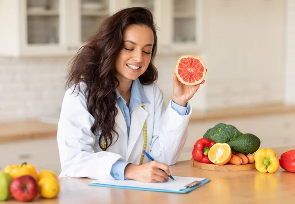 Happy caucasian woman doctor nutritionist making notes of diet plan at table with fruits, holding grapefruit, free space. Work, professional weight loss and health care