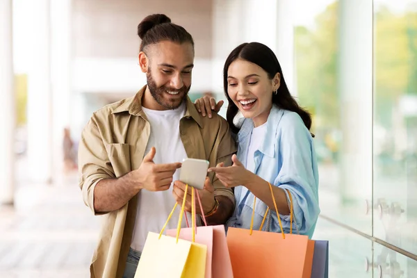 Smiling surprised young european man and woman with bags enjoy shopping and free time, look at phone in mall. Sale and shopaholics, discount season and app for buy, order, cashback