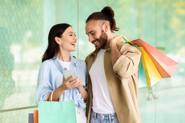 Laughing young european woman show smartphone to man with bags, recommend app, enjoy shopping and free time in mall. Shopaholics, discount and sale, cashback, blog and surprise
