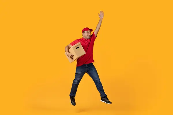 Delivery Service. Hispanic Young Courier Man In Red Uniform Jumping Holding Cardboard Box, Posing With Parcel In Mid Air, Raising Arm Over Yellow Studio Background, Full Length. Shipping Offer