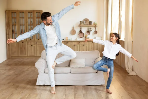 Joyful daddy and his daughter girl dancing to music and doing funny moves together while fooling at home. Father enjoying time with kid, having fun on weekend in living room indoors, full length