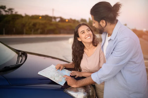 Enthusiastic latin couple reading map while having car trip together, looking for new destination, standing near the hood of car and talking, free space