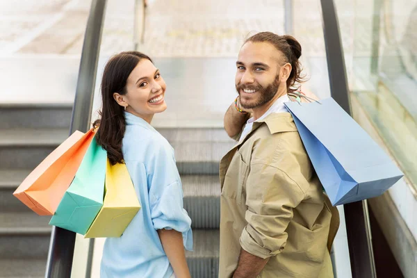 Glad young european couple with bags on escalator, enjoy shopping, free time in mall, ad and offer. Shopaholics have fun at sale, discount season, lifestyle love and relationships