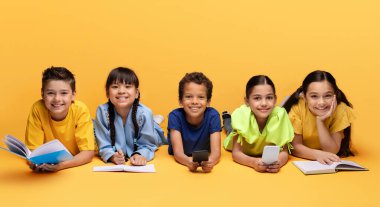 Çocuk yaşam tarzı ve eğlence, mutlu çocukluk konsepti. Neşeli çoklu etnik çeşitlilikteki çocuklar sarı arka planda izole bir şekilde yerde uzanıyorlar, kitap okuyorlar, ödev yapıyorlar, telefon kullanıyorlar, afiş asıyorlar.