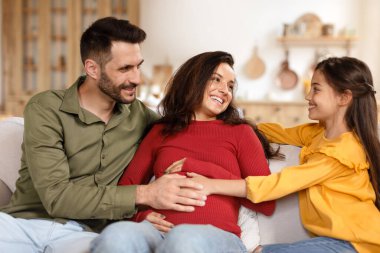 Aile İkinci Çocuğu Bekliyor. Anne karnına dokunan mutlu çocuk küçük kız kardeşini bekliyor, babasıyla koltukta oturuyor. Ebeveynler ve Kızları Bebek Beklentilerinin tadını çıkarıyorlar.