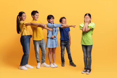 Okul çağında zorbalık. Sarı arka planda izole edilmiş ağlayan kızı işaret eden çok ırklı, küçük çocuklar. Zavallı çocuk sınıf arkadaşlarının alaylarından muzdarip.