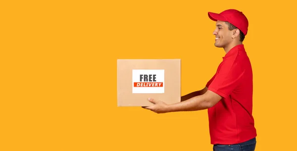 Free Delivery Offer. Hispanic Courier Guy Giving Carton Box Aside To Copy Space, Holding Carton Parcel On Yellow Studio Background. Side View Shot Of Deliveryman Shipping Purchase. Panorama