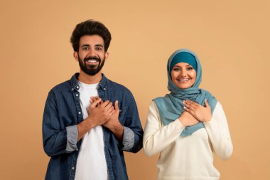 Minnettar müslüman çift ellerini göğsünde tutarak şükran ve takdirlerini sunuyorlar. Nazik Arap erkek ve kadın tesettürde iyi hisler bej rengi stüdyo arka planında duruyorlar.