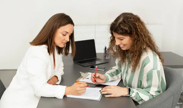 Hr manager lady signing job offer papers with employee woman sitting at office desk indoor. Side view shot of businesswoman consulting colleague, helping write business report. Career, employment