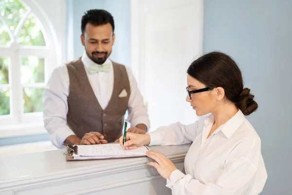 Young European Businesswoman And Receptionist Indian Man Filling Form And Signing Papers Checking At Hotel, Standing Near Reception Desk Indoor. Selective Focus On Lady Wearing Eyeglasses