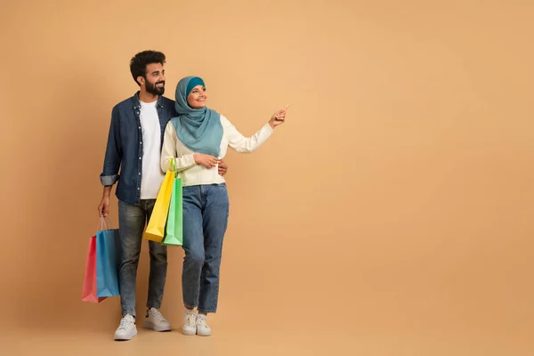 シーズンセールス ベージュスタジオの背景の上に立っている間 コピースペースで脇に指すショッピングバッグと幸せなムスリムカップル 笑顔のアラビアの配偶者が購入を楽しむ フル長さのショット — ストック写真