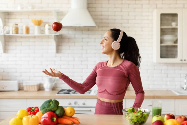 Easy Healthy Recipes. Cheerful Fit Lady With Headphones Throwing And Catching Red Apple Fruit In Kitchen, Cooking And Enjoying Dinner Preparation, Caring For Body And Weight Loss