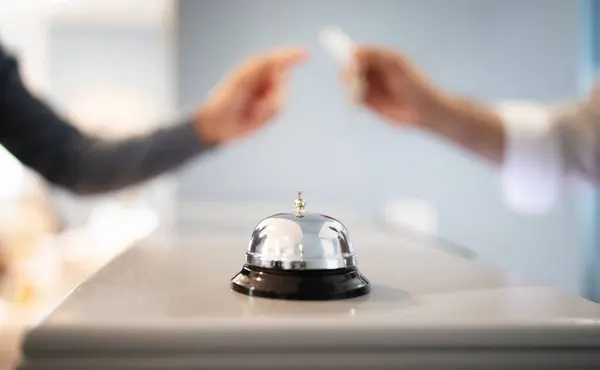 Call Bell Standing On Reception Counter At Hotel Lobby While Receptionist Giving Key To Tourist Man, Selective Focus, Closeup. Guest Checks In At Accommodation, Takes Room Keys From Hostel Staff