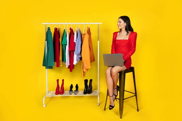 Online Personal Styling. Happy young woman sitting with laptop computer near clothing rack, female stylist looking at clothes and smiling, posing over yellow studio background, full length