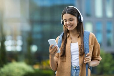 Üniversite kampüsünde kablosuz kulaklık ve akıllı telefon kullanarak sırt çantası taşıyan, uzun saçlı, hoş bir genç bayan. Çevrimiçi eğitim