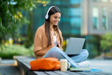 Gündelik öğrenci derslerine katılan güzel bir bayan, bankta oturuyor, bilgisayarını kullanıyor ve kablosuz kulaklık takıyor, kahve içiyor, fotokopi çekiyor. Çevrimiçi rota, webinar