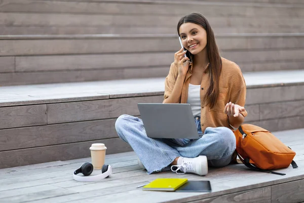 学生広告 コミュニケーションコンセプトのためのモバイルプラン 陽気で美しいブルネットの若い女性は 屋外に座って ノートパソコンを使用して テイクアウトコーヒーを飲んで 電話で話し 無料のスペースを見ます — ストック写真