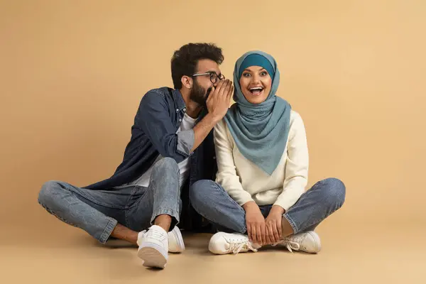 秘密を共有する 彼のガールフレンドの耳にささやく陽気なアラブ男 彼らはベージュスタジオの背景に座っている間 幸せなイスラム教徒のカップルは一緒にゴシップ ヒジャブのリスニングストーリーで興奮した女性 — ストック写真