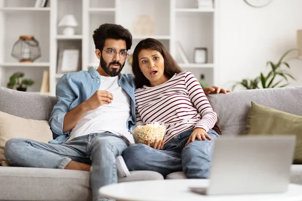 Frightened young eastern couple man and woman sitting on couch in cozy living-room, eating popcorn, watching shocking content on TV, lovers spending weekend together at home