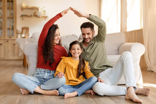 Real estate offer. Young middle eastern dad, mom and daughter sitting under symbolic roof, dreaming of new home together, posing on floor in modern living room interior. Housing for young family