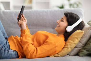 Rahat, genç Hintli kadın kanepede uzanıyor, evde kablosuz kulaklık ve cep telefonu kullanıyor. En sevdiği podcast 'i izleyen mutlu Hintli kadın, çevrimiçi müzik dinliyor, video görüşmesi yapıyor, yan görüş yapıyor.