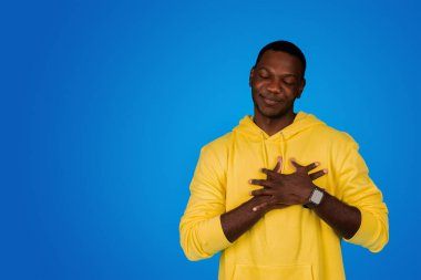 Kapalı gözlü, pozitif genç bir adam göğüs göğüse, teşekkür jesti yapıyor, mavi arka planda, stüdyoda izole edilmiş. Minnettarlık, aşk işareti, insani duygular, reklam ve teklif, yaşam tarzı