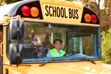 Portrait Of Happy Black Female Driver Driving Yellow School Bus, Beautiful Young African American Lady In Reflecting Vest Looking Through Window, Holding Steering Wheel An Smiling At Camera clipart