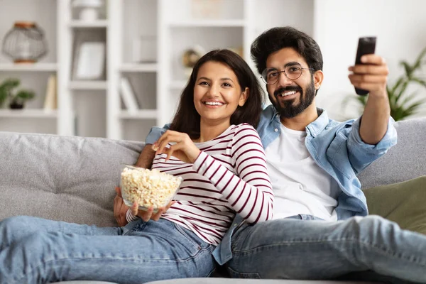 Cinema time. Portrait of happy relaxed beautiful millennial indian couple sitting on couch in living room, eating popcorn, bonding, watching TV, choosing movie, enjoying evening at home, copy space