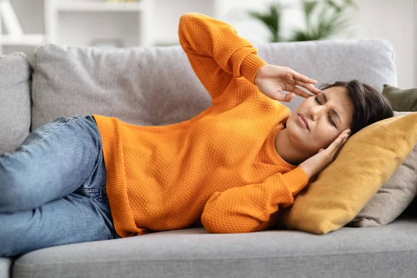 Weak sick millennial indian woman lying on couch at living room feel dizzy migraine headache suffer from sleep problem disorder. Young eastern lady wake up after nap lie on side, rubbing temples