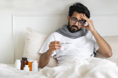 Yatakta oturan ve termometreyi tutan hasta Hintli adam vücut ısısını ölçen genç, grip belirtileri gösteren ve hayal kırıklığı içinde alınına dokunan, boş alan olan genç adam.